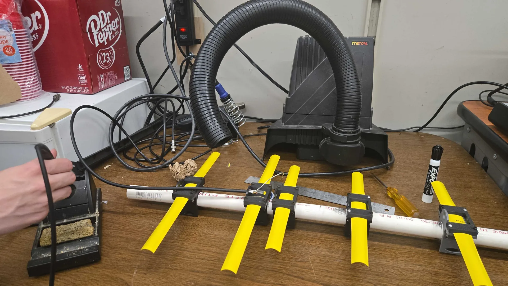 Workbench view of the 70 cm tape-measure Yagi feedpoint during soldering before final assembly, with a soldering iron, fume extractor, PVC boom, and yellow tape elements visible